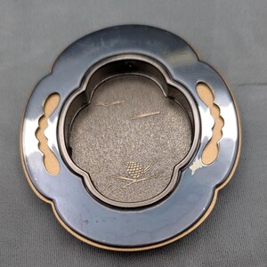 Peut inclure: Un dessous de verre décoratif en métal argenté avec un design unique à bord festonné. Le dessous de verre a une bordure beige clair et un intérieur texturé gris. Un motif de pomme de pin et de branche doré est au centre.