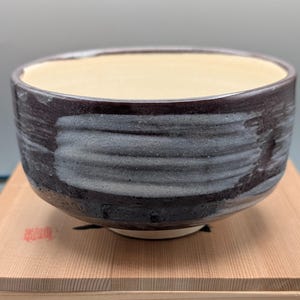 May include: A handcrafted ceramic bowl with a dark, textured exterior and a light beige interior. The bowl has a wide, rounded shape with visible brushstrokes in shades of gray and black. It sits on a wooden surface, showcasing its artisanal quality.