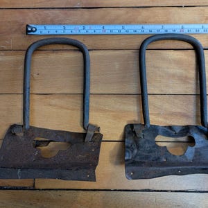 Puede incluir: Dos soportes metálicos oxidados con una parte superior curva y una base rectangular. Los soportes tienen un agujero en el centro de la base.