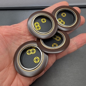 May include: Three circular brown buttons with a recessed black center. Each button features a metallic gold rim with a fine textured edge and a graphic pattern of yellow circular symbols printed on the black interior surface of the button.