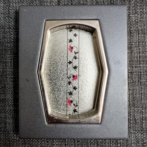 May include: A rectangular gray picture frame with a silver interior. The frame holds a decorative panel with a floral design of pink flowers and black leaves. The panel has a silver, textured background.
