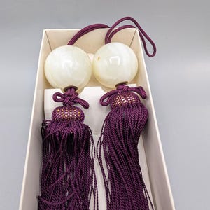May include: Two decorative curtain tiebacks featuring large, smooth, off-white spherical stone finials attached to deep purple, multi-strand silk tassels. Each tassel is secured with a knotted cord and a gold-flecked, dark purple decorative cap.