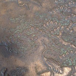 K&ouml;nnte beinhalten: Ein rundes, bronzenes Tablett mit einem gepr&auml;gten Design aus Kranichen und einer Kiefer. Das Tablett hat eine strukturierte Oberfl&auml;che und eine gr&uuml;ne Patina.