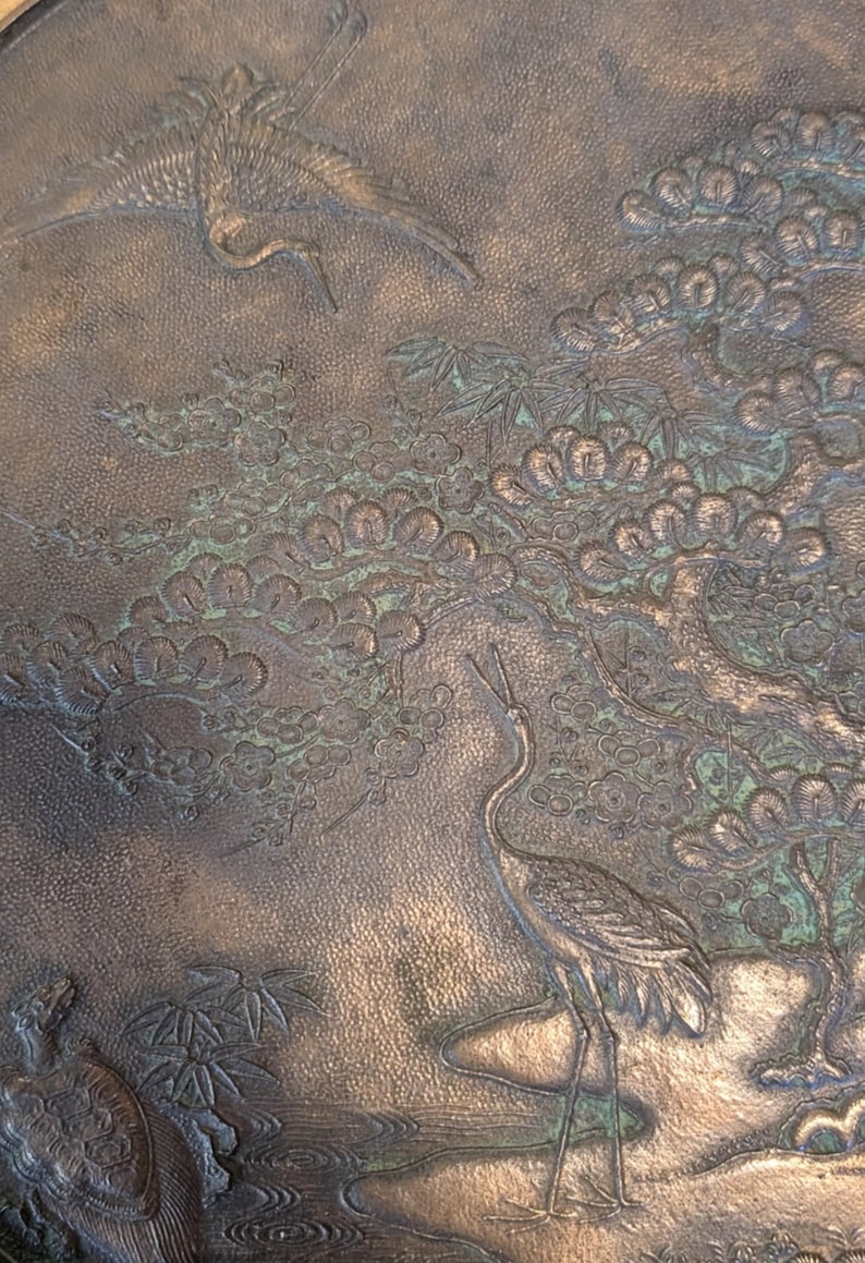 K&ouml;nnte beinhalten: Ein rundes, bronzenes Tablett mit einem gepr&auml;gten Design aus Kranichen und einer Kiefer. Das Tablett hat eine strukturierte Oberfl&auml;che und eine gr&uuml;ne Patina.
