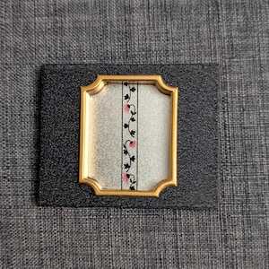 以下が含まれることがあります： 金色のフレームと黒いテクスチャの背景を持つ装飾的な長方形のオブジェクト。中央には、黒とピンクの垂直の花柄のデザインが施された銀色のパネルがあります。