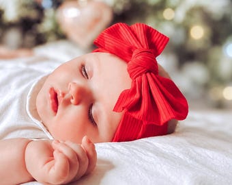 May include: A sleeping infant wearing a bright red ribbed fabric headband featuring a large, prominent knotted bow detail. The headband is positioned across the forehead, contrasting with the white clothing and soft, neutral textures.