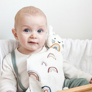 May include: A baby is holding a white muslin baby blanket with a rainbow pattern and a plush bunny toy with a rainbow and a smiling face.