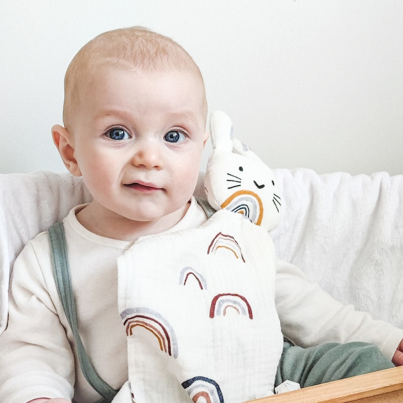 May include: A baby is holding a white muslin baby blanket with a rainbow pattern and a plush bunny toy with a rainbow and a smiling face.