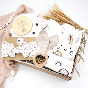 May include: A gift box filled with baby items, including a white onesie with the text "hello world" on a wooden circle, a white headband with rainbow designs, a wooden teether with the text "mama bear & cubs", and a white bunny-shaped plush toy with rainbow designs.