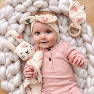 May include: A cream-colored headband with a knotted top and pink and teal heart print, a matching cream-colored bunny-shaped security blanket with heart accents, and a wooden teething ring with cream-colored bunny ears featuring heart prints.