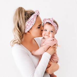 May include: A matching set of two headbands featuring a white fabric base with a small red floral pattern. Both headbands are designed with a knotted top detail. One headband is sized for an adult, and the other is sized for an infant.