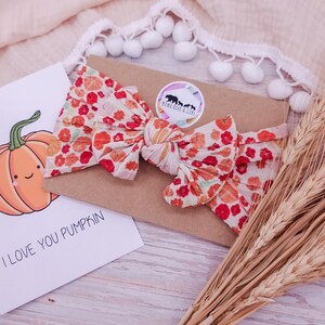 May include: A fabric headband featuring a knotted bow design with a repeating pattern of orange pumpkins and red flowers. The headband is attached to a rectangular brown card with a circular sticker that reads Mama Bear & Cubs.