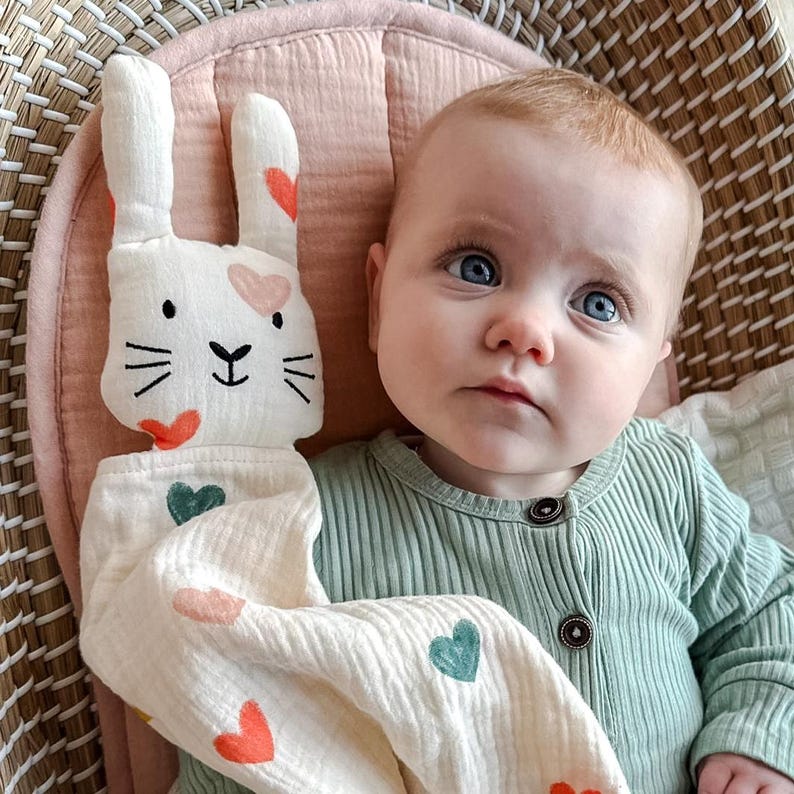 May include: A baby is lying in a wicker basket with a white muslin bunny lovey. The lovey has pink, green, and orange hearts on it. The baby is wearing a green ribbed onesie.