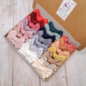May include: A collection of ribbed fabric hair bows in a variety of colours including white, cream, tan, yellow, orange, pink, red, blue, and grey, arranged in a cardboard gift box with a white sticker featuring a bear logo and text.