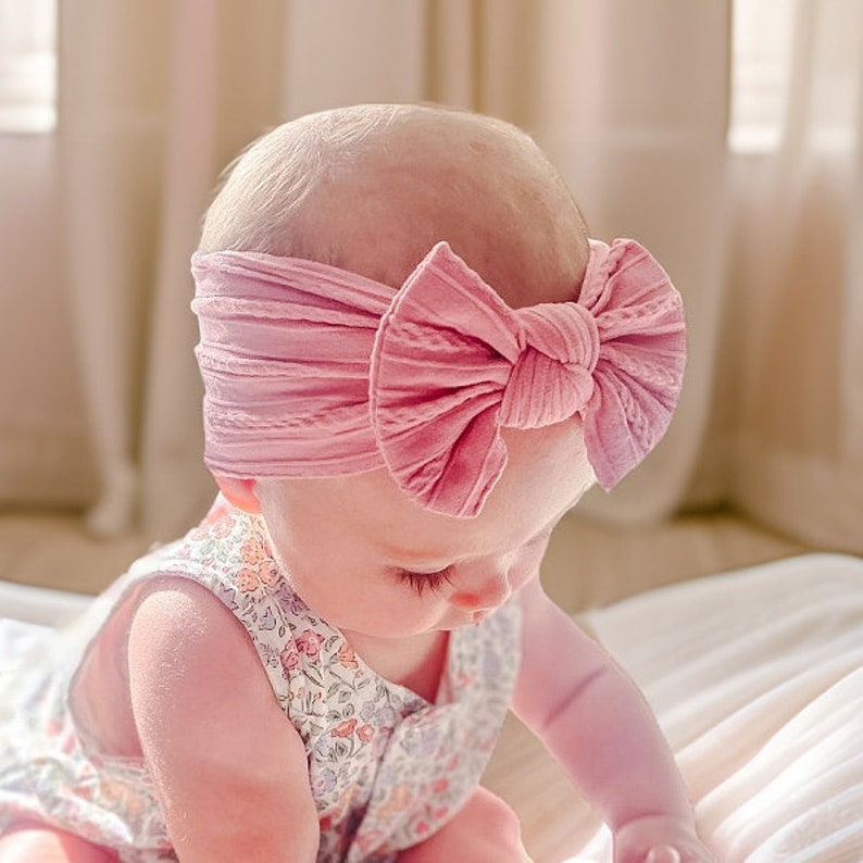 May include: A baby wearing a pink ribbed headband with a large knotted bow. The headband is made of soft, stretchy fabric and is perfect for newborns and toddlers.