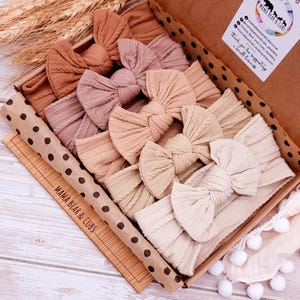 K&ouml;nnte beinhalten: Ein Set mit sechs geknoten Stirnb&auml;ndern in verschiedenen Braunt&ouml;nen, Beige und Tan, angeordnet in einer Schachtel mit Tupfenmuster und dem Text "Mama Bear & Cubs".