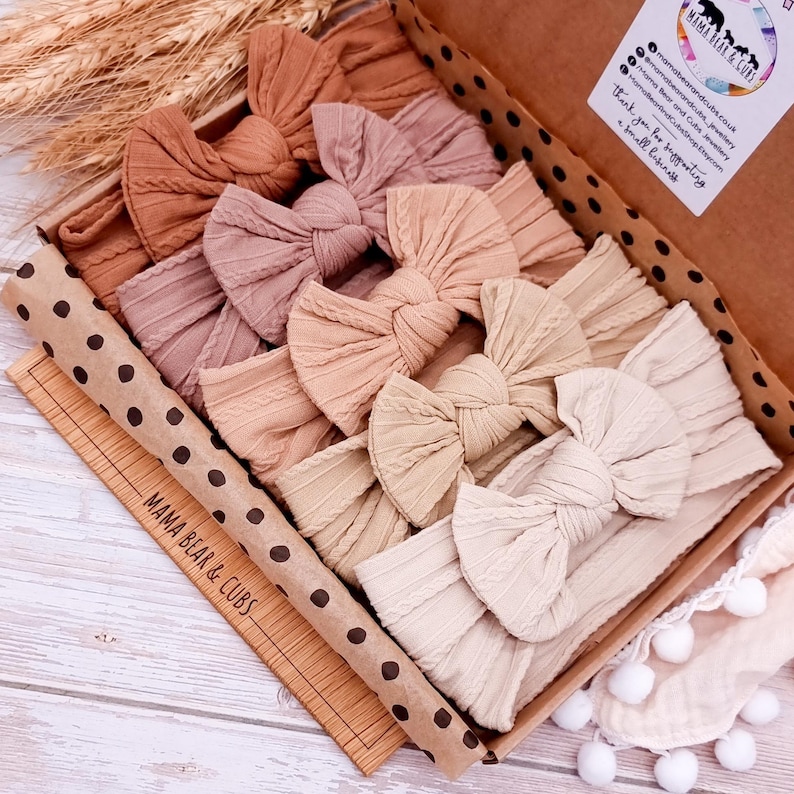 K&ouml;nnte beinhalten: Ein Set mit sechs geknoten Stirnb&auml;ndern in verschiedenen Braunt&ouml;nen, Beige und Tan, angeordnet in einer Schachtel mit Tupfenmuster und dem Text "Mama Bear & Cubs".