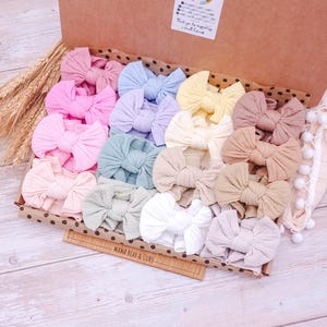 May include: A collection of ribbed fabric hair bows in various pastel and neutral colors, including pink, blue, yellow, lavender, and beige, arranged inside a cardboard box labeled MAMA BEAR & CUBS with a thank you note attached to the lid.