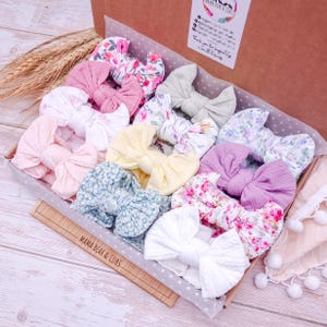May include: A cardboard box containing a collection of fabric hair bows in various colours and patterns, including solid pink, yellow, white, and purple, alongside floral prints in blue, pink, and green, with a Mama Bear & Cubs label attached.