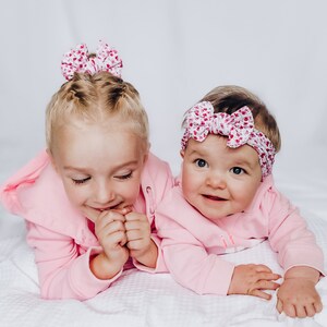 May include: Two light pink hooded sweatshirts, one worn by a child with a floral bow hair accessory and the other by an infant wearing a matching floral headband. Both garments feature long sleeves and a soft, solid light pink fabric construction.