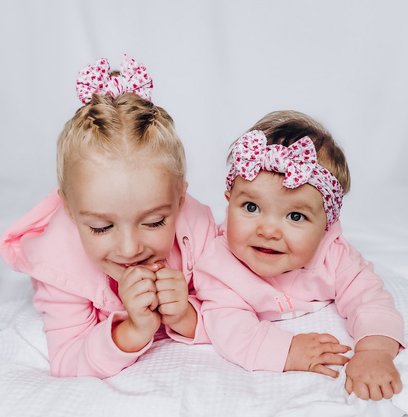 May include: Two light pink hooded sweatshirts, one worn by a child with a floral bow hair accessory and the other by an infant wearing a matching floral headband. Both garments feature long sleeves and a soft, solid light pink fabric construction.