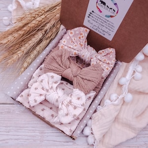 May include: A cardboard gift box containing three fabric headbands with knotted bows. One headband features a white daisy print on a pink background, one is a solid dusty rose cable knit, and one has a small floral pattern on a light pink base.