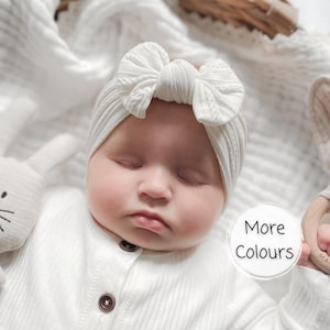 May include: A white ribbed baby headband featuring a large, centred fabric bow. The headband is made of a soft, textured material and is worn by an infant. A circular graphic overlay on the right side contains the black text More Colours.