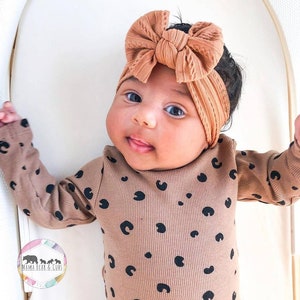May include: A long-sleeved ribbed bodysuit in a tan color featuring a black leopard spot pattern, paired with a matching tan ribbed headband that has a large, knotted bow detail on the front. The garment includes a small circular logo.