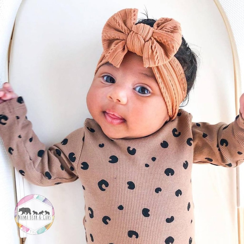 May include: A long-sleeved ribbed bodysuit in a tan color featuring a black leopard spot pattern, paired with a matching tan ribbed headband that has a large, knotted bow detail on the front. The garment includes a small circular logo.