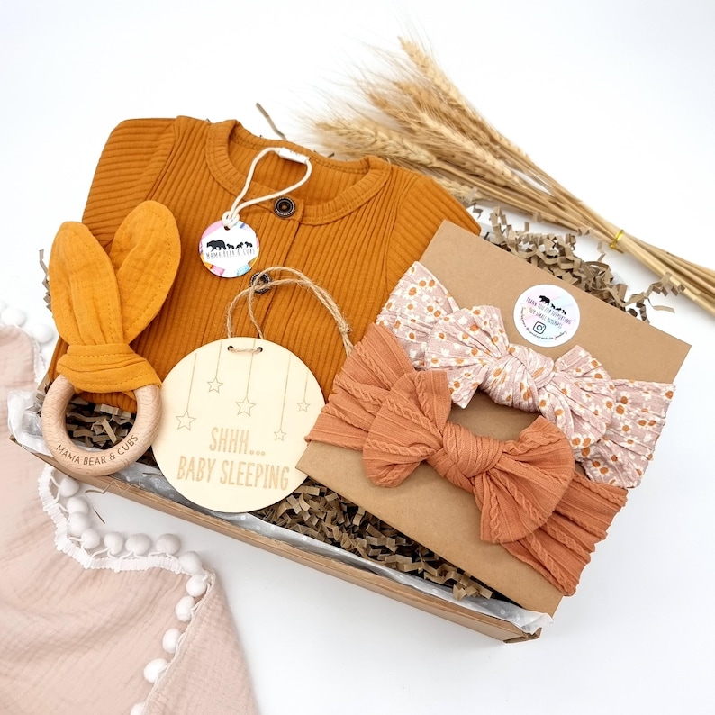 Puede incluir: Un juego de regalo para beb&eacute;s en una caja de madera. El juego incluye una rebeca de color mostaza, un mordedor con forma de orejas de conejo y tres diademas con lazos. Se incluye un letrero de madera con el texto "Shhh... Baby Sleeping".