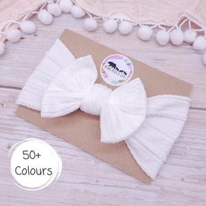 May include: A white ribbed fabric headband featuring a large, centred bow. The headband is attached to a rectangular brown card with a circular sticker displaying the text Mama Bear & Curls. A separate circular graphic shows the text 50+ Colours.