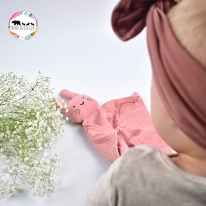 May include: A pink bunny-shaped security blanket with a smiling face. The blanket is made of soft, textured fabric and is perfect for cuddling.
