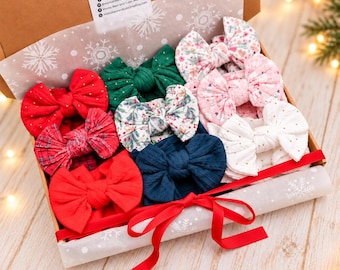 May include: A cardboard gift box containing a collection of fabric hair bows in various colours and patterns, including solid red, dark green, navy blue, white with gold dots, floral prints, and plaid, tied with a red ribbon and snowflake tissue paper.