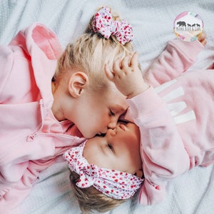 May include: Two children wearing matching light pink zip-up hoodies and floral print hair accessories. One child wears a floral bow in a high bun, while the other wears a floral headband. A logo in the corner reads Mama Bear & Cubs.