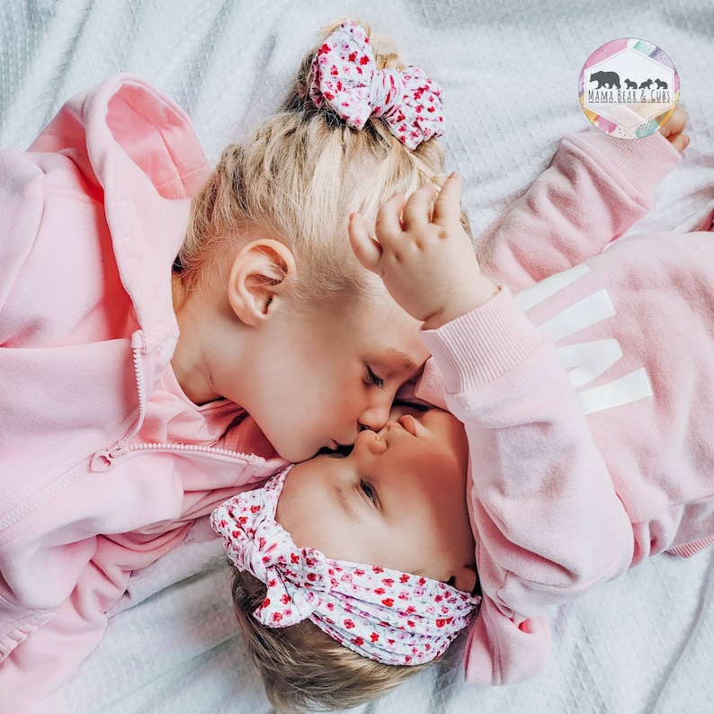May include: Two children wearing matching light pink zip-up hoodies and floral print hair accessories. One child wears a floral bow in a high bun, while the other wears a floral headband. A logo in the corner reads Mama Bear & Cubs.