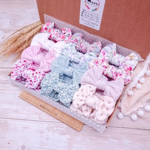 May include: A cardboard box containing a collection of fabric hair bows in various floral and solid pastel patterns. The bows feature knotted centres and are arranged in rows. A white label with the text Mama Bear & Cubs is attached to the box lid.