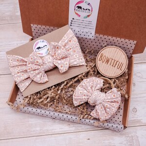 May include: A light pink ribbed fabric headband and a matching hair bow, both featuring a small white daisy print. The items are packaged in a cardboard box with brown paper filler, a circular wooden tag engraved with the word BOWTIFUL, and a logo sticker.