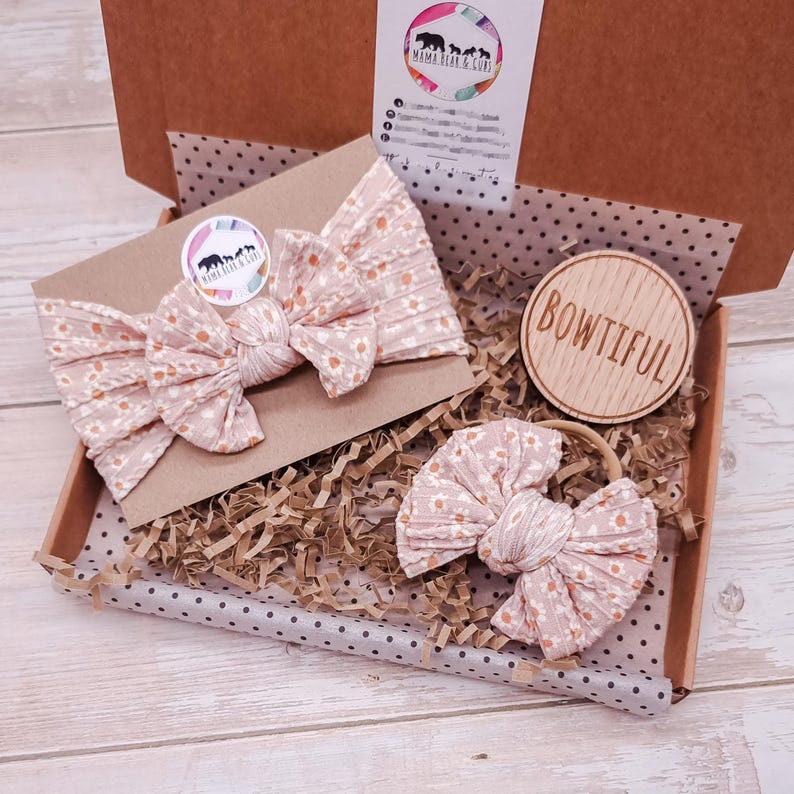 May include: A light pink ribbed fabric headband and a matching hair bow, both featuring a small white daisy print. The items are packaged in a cardboard box with brown paper filler, a circular wooden tag engraved with the word BOWTIFUL, and a logo sticker.