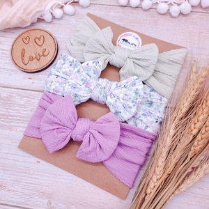 May include: Three fabric headbands with bows attached to a rectangular card. The top headband is sage green with a cable knit texture. The middle headband features a floral pattern with purple and green tones. The bottom headband is solid lavender.