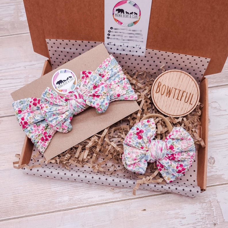 May include: A cardboard gift box containing a floral print fabric headband and a matching floral print fabric bow on a nylon band. The box includes a circular wooden tag engraved with the word BOWTIFUL and a branded card with the text MAMA BEAR & CUBS.