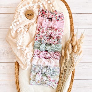 May include: A collection of ten baby headbands featuring various floral and solid patterns in shades of pink, green, and white. Each headband is designed with a central knotted bow detail and is arranged in a vertical stack on a cream fabric.