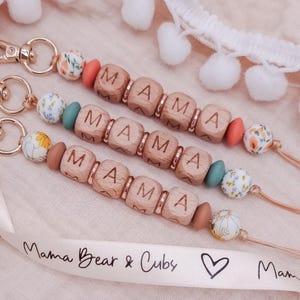 May include: Three wooden keychains with the word "MAMA" spelled out in individual wooden beads. Each keychain has a different coloured bead and floral pattern. The keychains are displayed on a white surface with a white ribbon that says "Mama Bear & Cubs" and a heart.