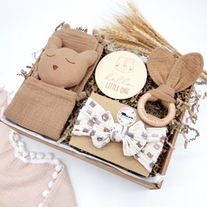 May include: A brown gift box filled with baby items. The box contains a brown muslin swaddle, a brown and white headband with a bear print, a wooden teether with the text "Mama Bear & Cubs", a brown plush toy with a sleeping face, and a wooden round sign with the text "hello little one".