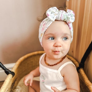 May include: A baby wearing a white headband with a floral pattern and a brown amber necklace. The headband has a large bow in the centre. The baby is sitting in a wicker basket.