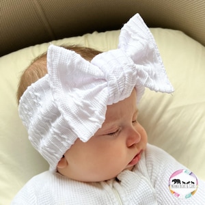 May include: A white knitted headband with a large bow on top. The headband is worn by a baby sleeping on a white surface.