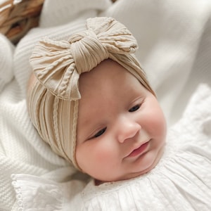 Puede incluir: Una diadema de nudo de color beige con textura de canalé y un gran lazo en la parte superior. La diadema la lleva un bebé.