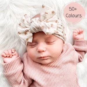May include: A baby headband featuring a large, knotted bow design in an off-white fabric with a subtle tan floral pattern. The headband is worn by a sleeping infant dressed in a long-sleeved, ribbed, dusty pink shirt. A circular graphic states 50+ Colours.