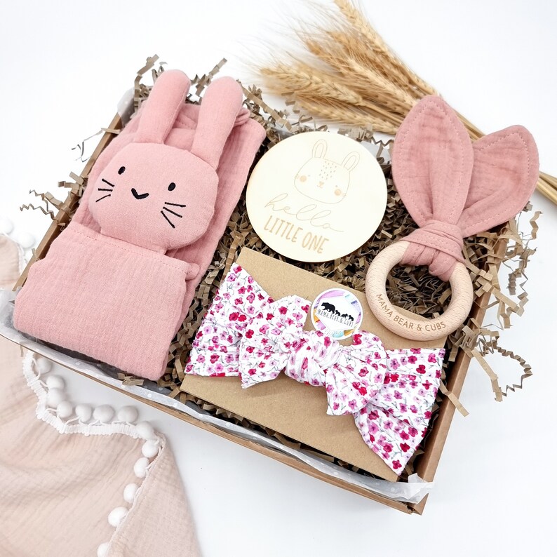 May include: A pink and white baby gift box with a pink bunny rattle, a pink bunny headband, a pink and white floral headband, a wooden bunny plaque that says "hello little one", and a wooden ring with the text "mama bear & cubs".
