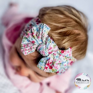 May include: A textured fabric headband featuring a knotted bow design with a colorful floral pattern of pink, blue, and green blossoms on a white background. The accessory is worn around the head, showcasing a gathered, soft material construction.