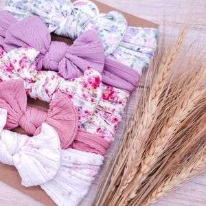 May include: A collection of five fabric headbands featuring knotted bows, arranged on a rectangular card. The headbands include solid mauve and pink cable-knit textures alongside white fabric with floral patterns in shades of pink, purple, and green.